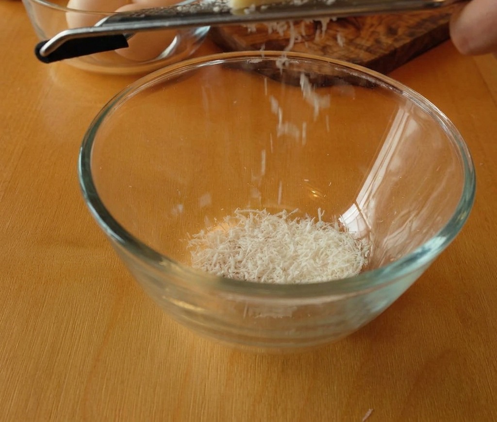 A block of Pecorino Romano being grated into a glass bowl, creating a pile of fine white cheese shavings.