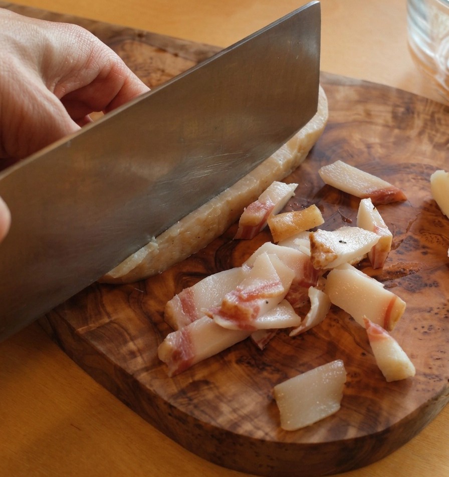 A sharp knife trimming the thick, dark skin off a slab of guanciale on a rustic wooden board.