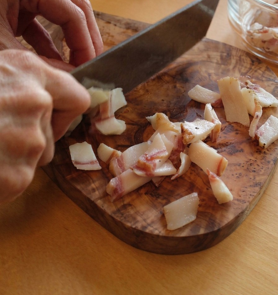 Slicing guanciale into small rectangular strips on a cutting board, showing the layers of white fat and pink meat.