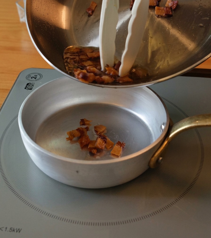 Pouring golden-brown, crispy guanciale bits and rendered liquid fat from a large pan into a small prep bowl.