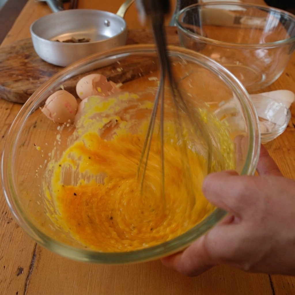 A hand using a whisk to blend bright yellow egg yolks and grated cheese in a glass bowl while hot water is added.