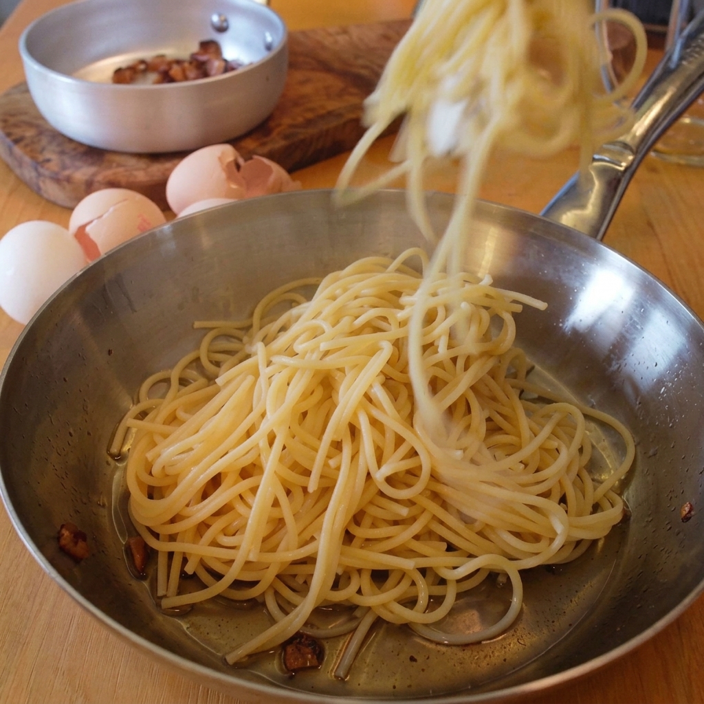 Tongs dropping freshly cooked spaghetti into a stainless steel pan coated with melted guanciale fat.