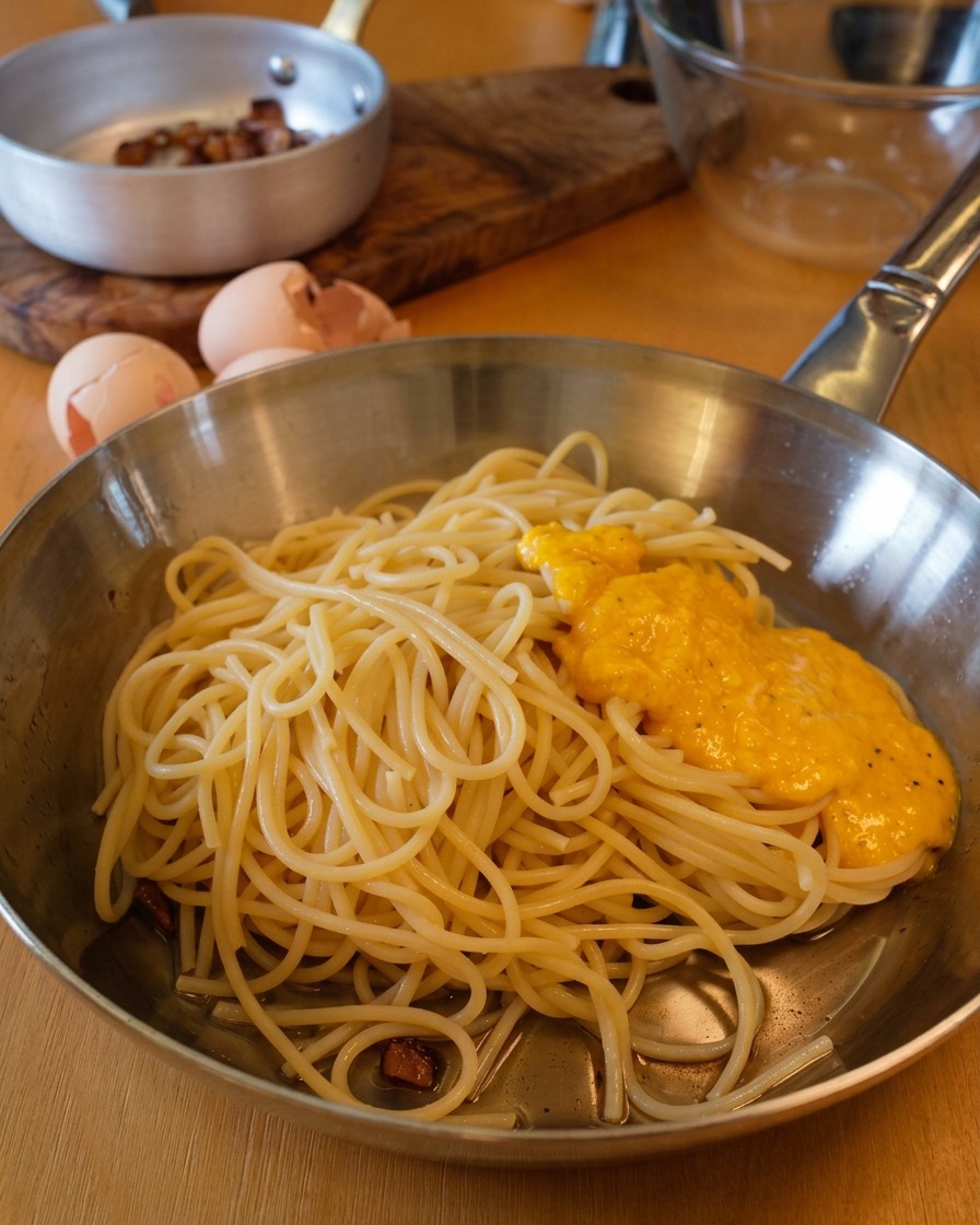 A thick, golden egg and cheese sauce resting on top of a mound of spaghetti in a stainless steel pan.