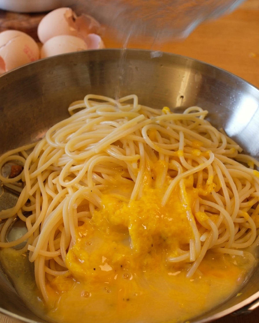 Spaghetti in a stainless steel pan being coated with a thick, yellow egg yolk sauce as a stream of pasta water is poured over it.