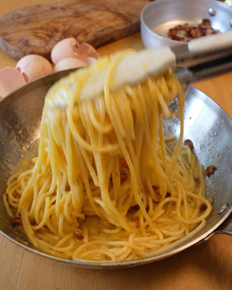 Tossing spaghetti in a metal pan with tongs to create a creamy Carbonara sauce.