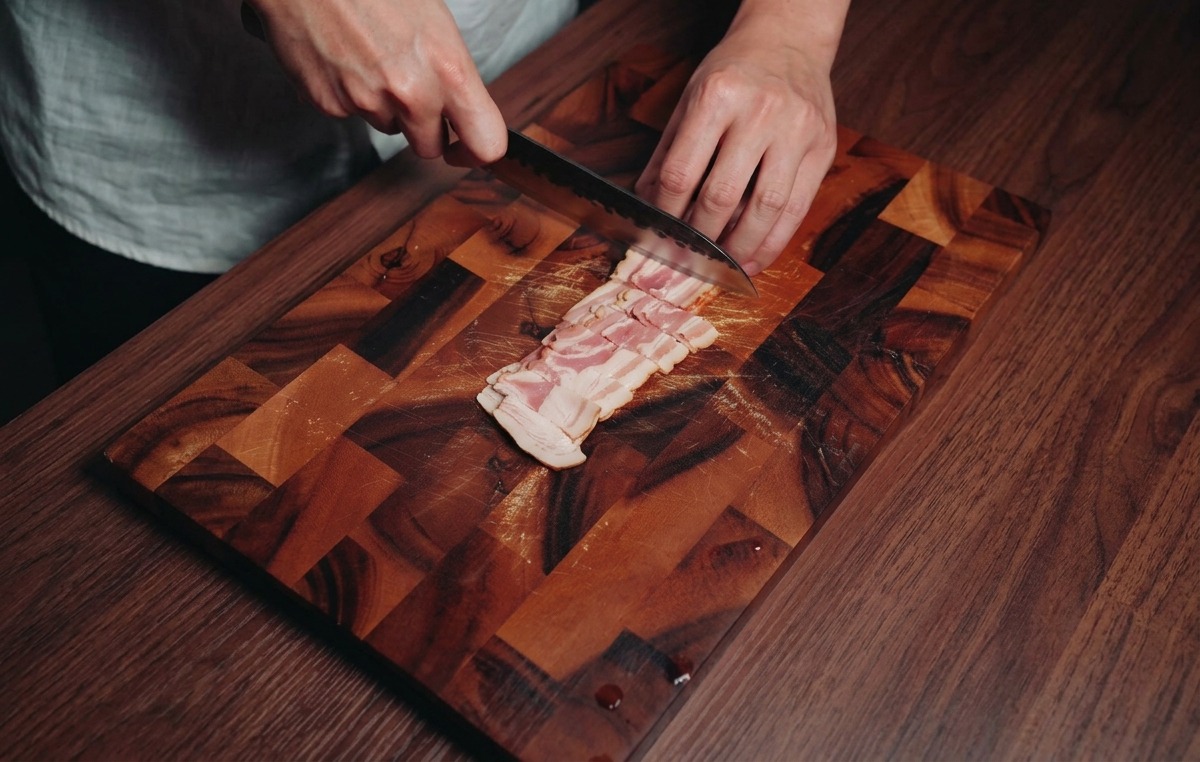 A person using a chefs knife to cut strips of raw marbled bacon into small rectangular pieces on a dark wood board.