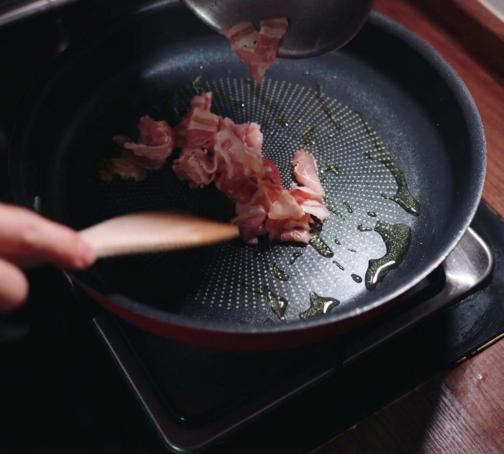 Sliced bacon pieces being poured from a bowl into a black frying pan containing a small amount of oil.