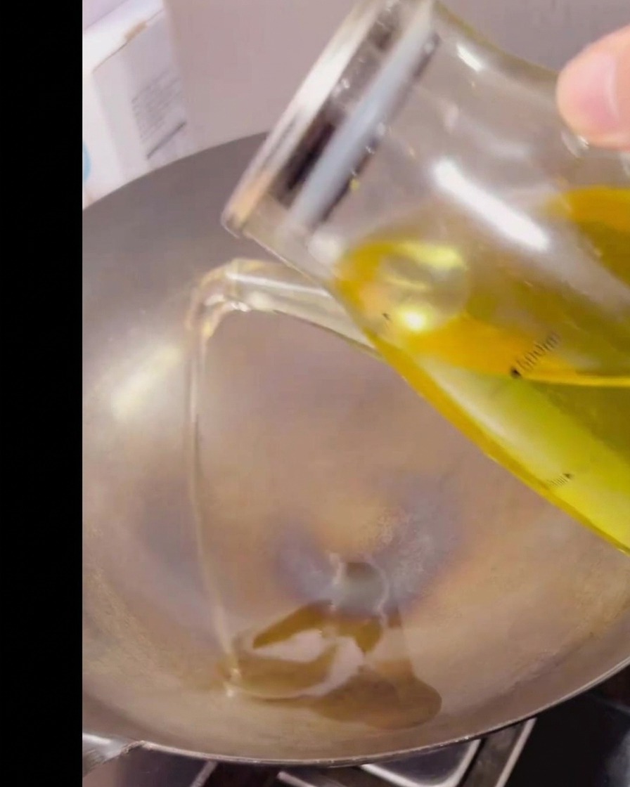 Clear cooking oil being poured from a glass measuring pitcher into a hot, empty metal wok.