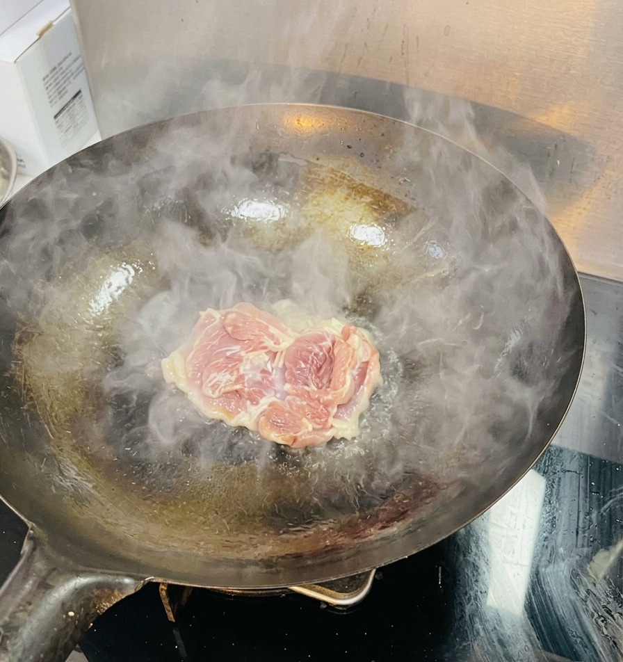 A single raw chicken thigh being placed into a hot wok with visible steam and shimmering oil.