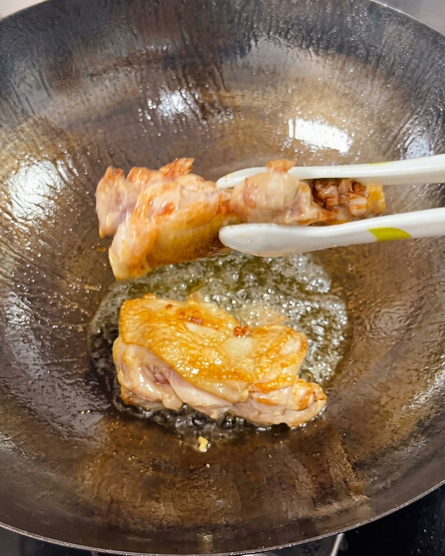 Tongs flipping a seared chicken thigh in a wok, showing the crispy, golden-brown cooked side.