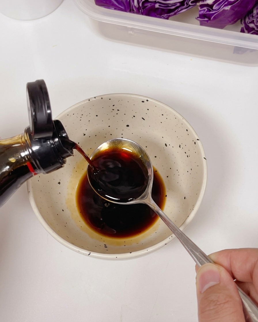 Light soy sauce being poured from a bottle into a spoon over a bowl containing dark sauce and seasonings.