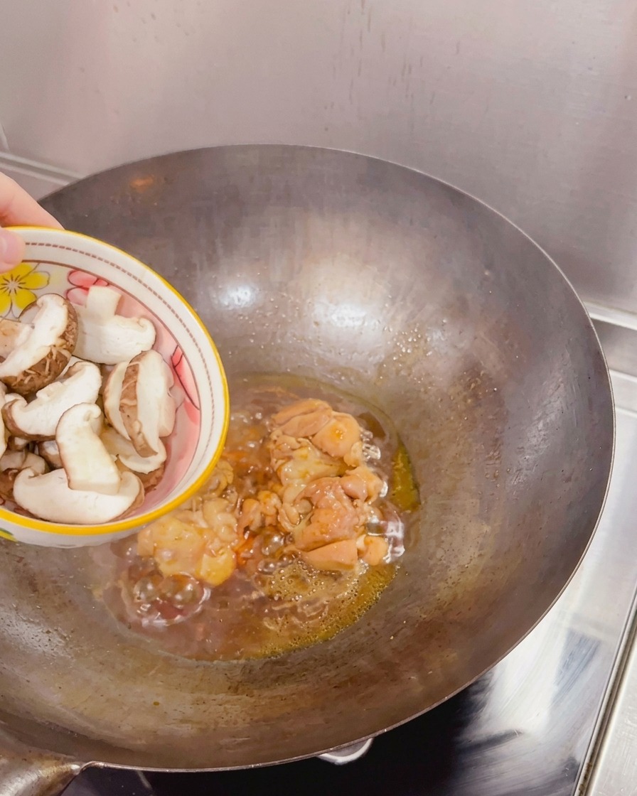 Sliced fresh shiitake mushrooms being added from a bowl into a wok containing seared chicken and simmering sauce.