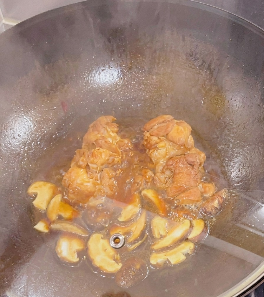 Chicken thighs and sliced shiitake mushrooms simmering in a dark sauce inside a wok under a glass lid.