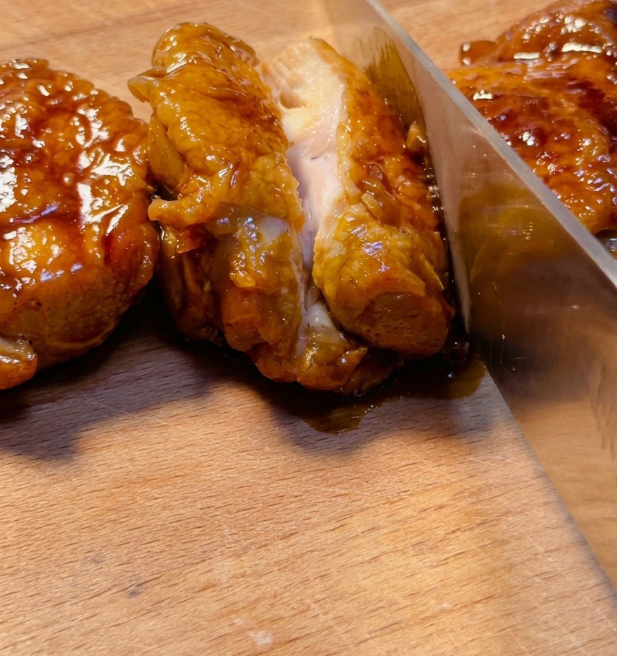 A sharp knife slicing through a deeply glazed teriyaki chicken thigh on a wooden board, showing the juicy meat inside.
