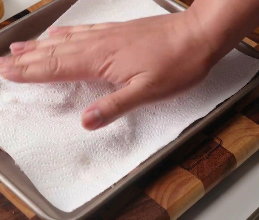 A hand using a white paper towel to pat dry raw chicken thighs laid out on a metal baking tray.
