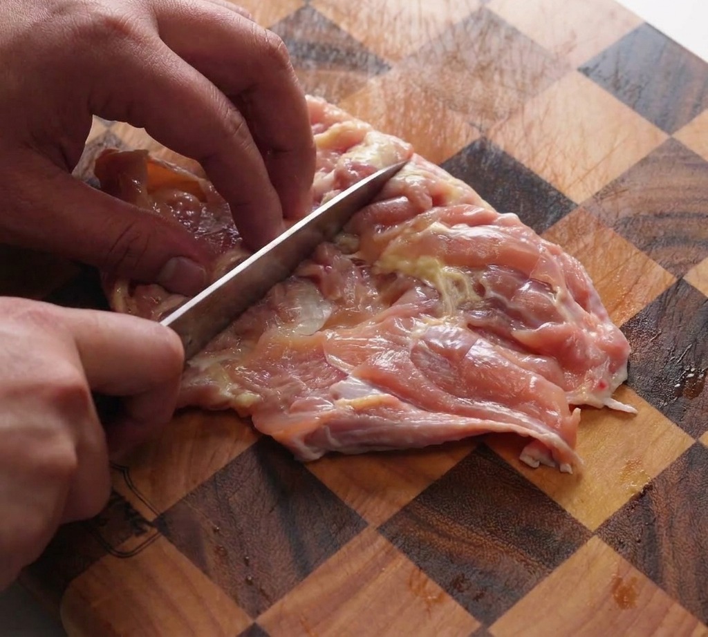 A hand using the back of a chefs knife to pound deboned chicken thighs on a checkered wooden cutting board.
