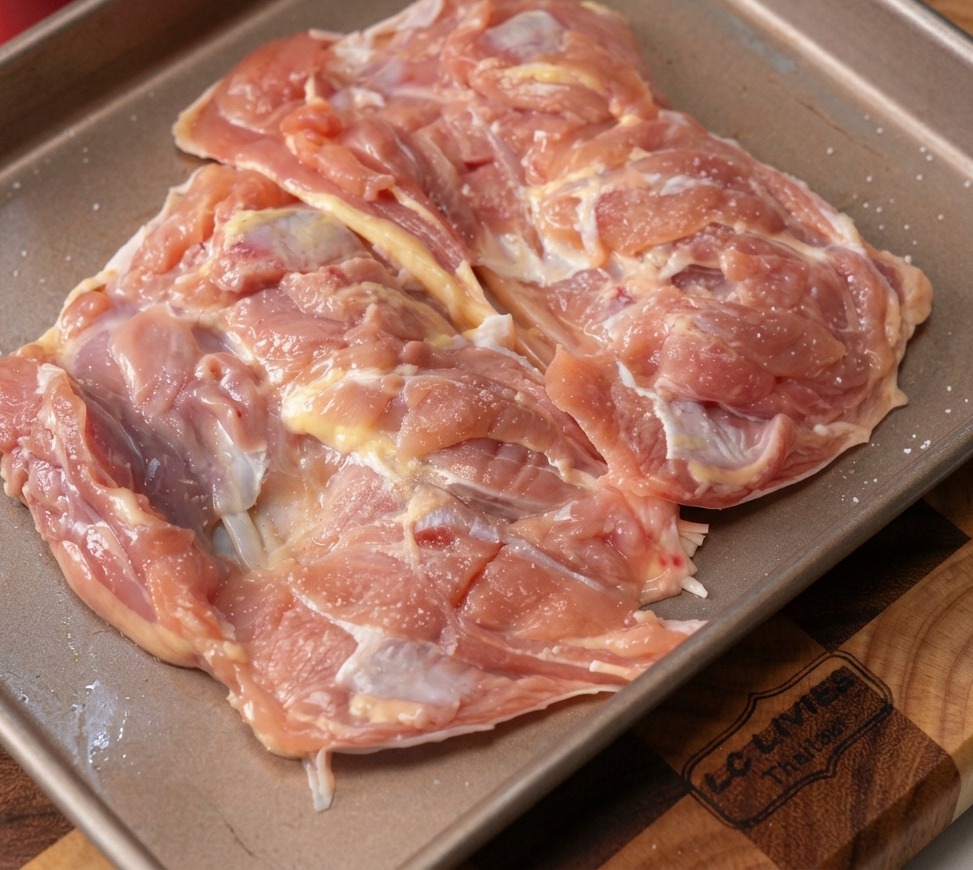 Raw deboned chicken thighs on a metal baking tray sprinkled with minced garlic and black pepper.