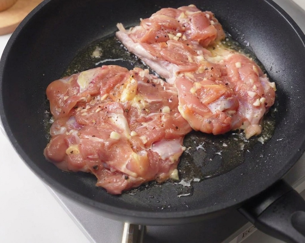 Two marinated chicken thighs being seared skin-side down in a dark non-stick frying pan.