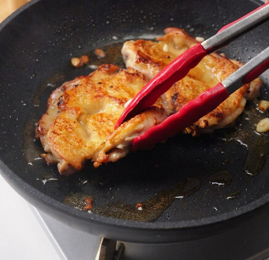 A hand pouring dark teriyaki sauce from a glass bowl over the seared chicken thighs in a pan.