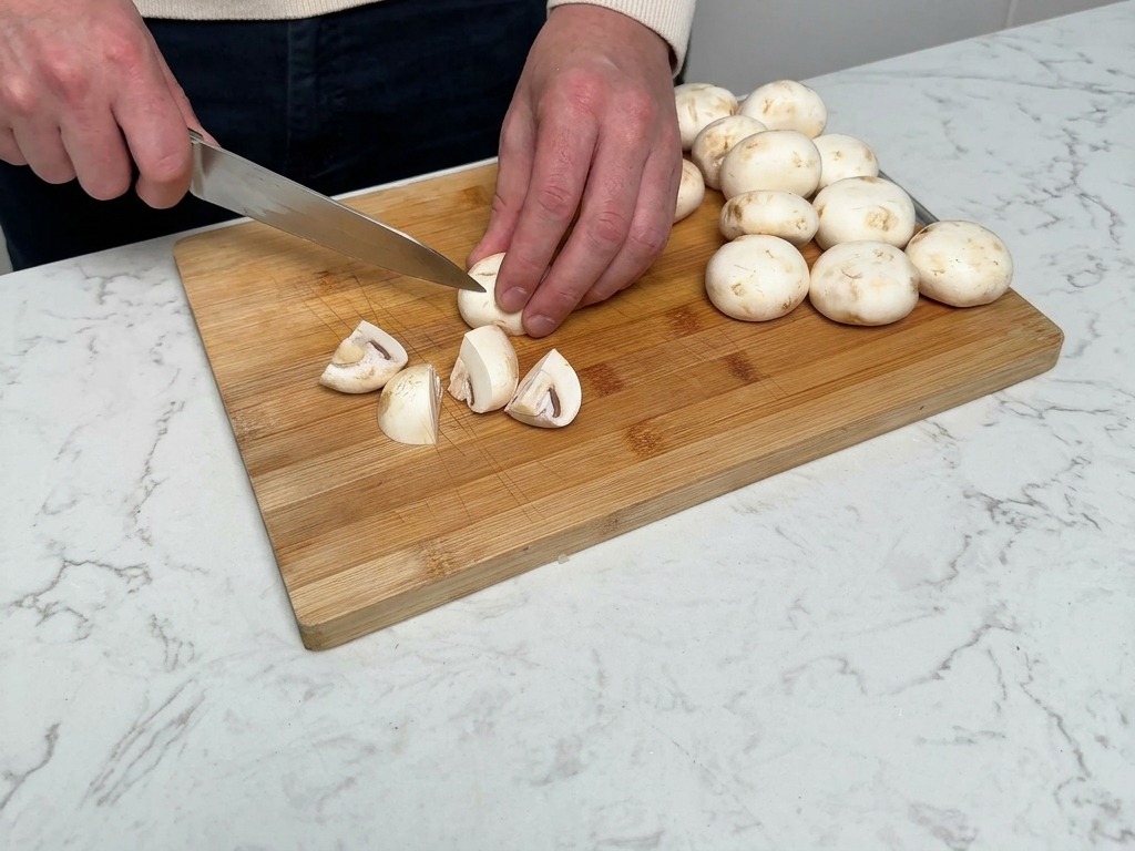 Hands using a chefs knife to cut white button mushrooms into quarters on a wooden chopping board.