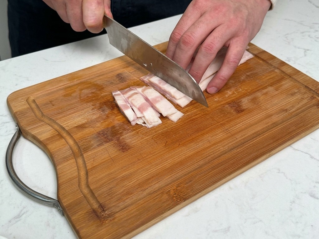 A person slicing thick strips of bacon on a wooden cutting board with a knife.