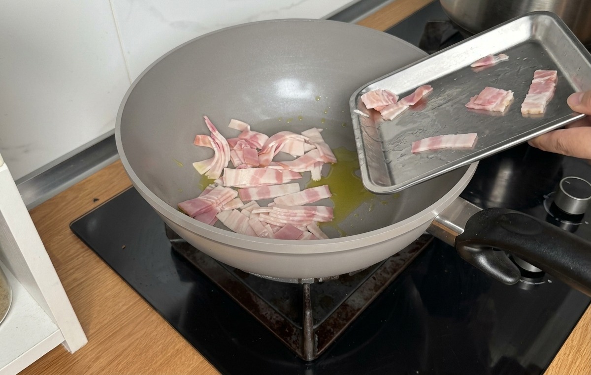 Sliding raw bacon strips from a small metal tray into a grey pan coated with olive oil.