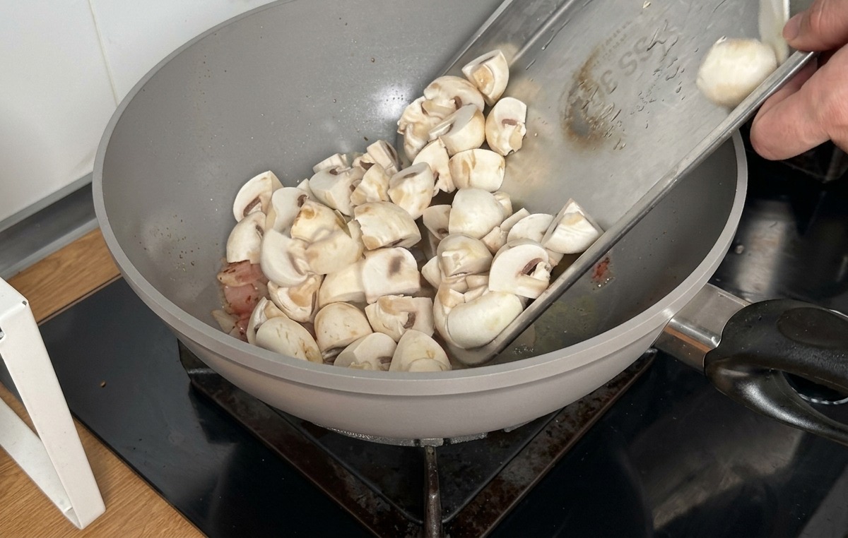 Pouring quartered white button mushrooms from a metal tray into a wok containing cooked bacon.