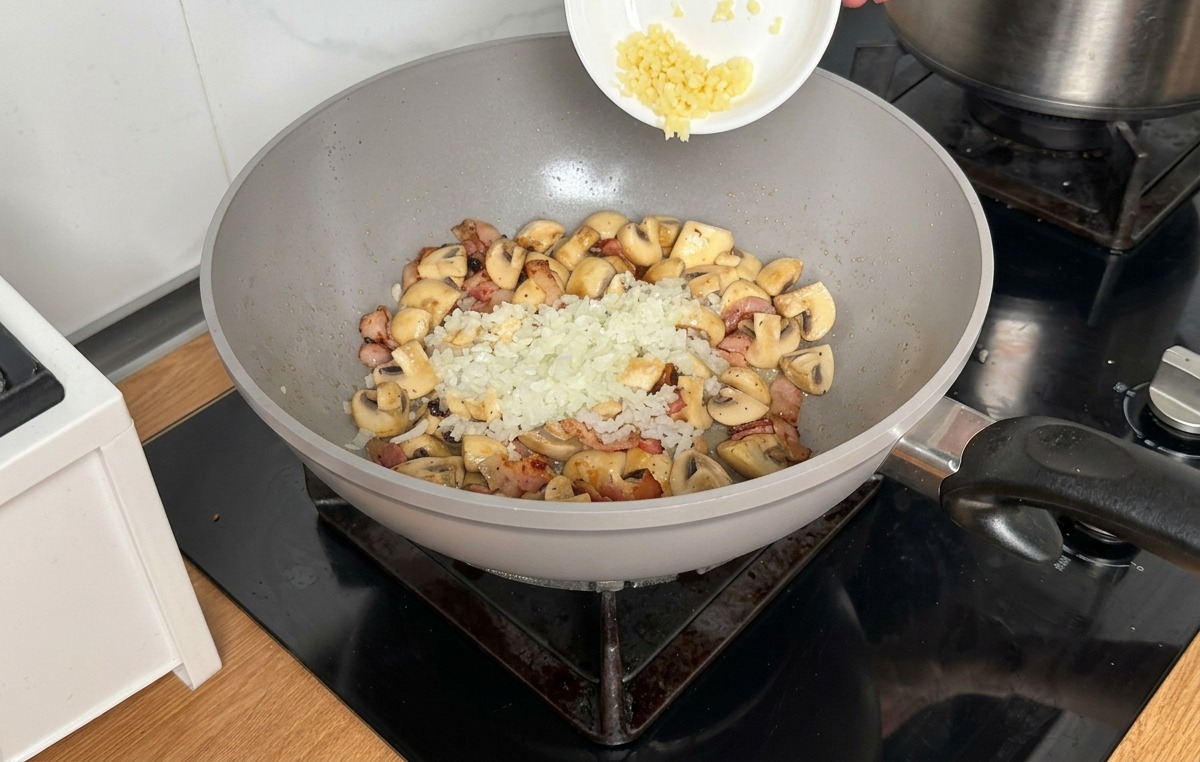 Pouring finely minced onions and garlic from a small white bowl over the cooked mushrooms and bacon in a wok.