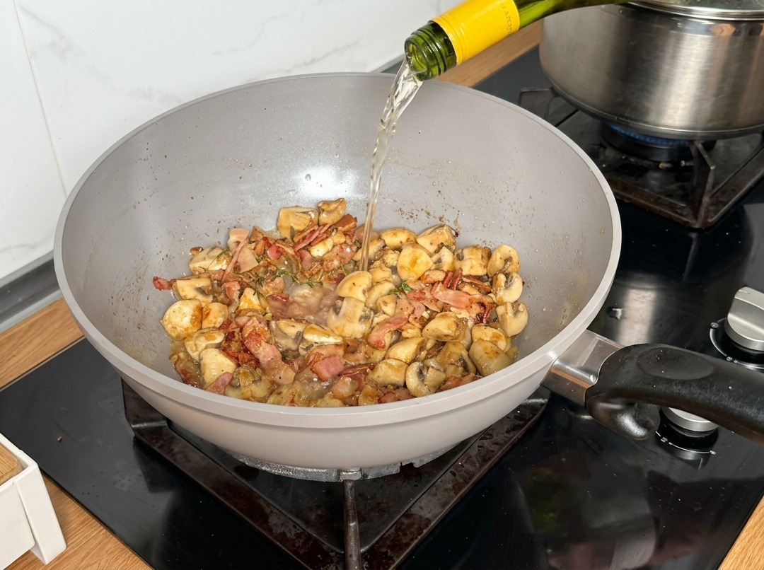 White wine being poured from a bottle into a pan containing cooked mushrooms and bacon.