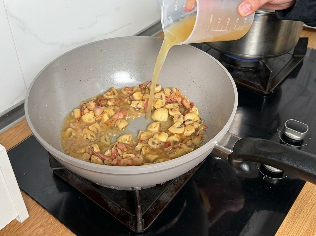 Chicken broth being poured from a plastic measuring cup into the mushroom and bacon mixture.