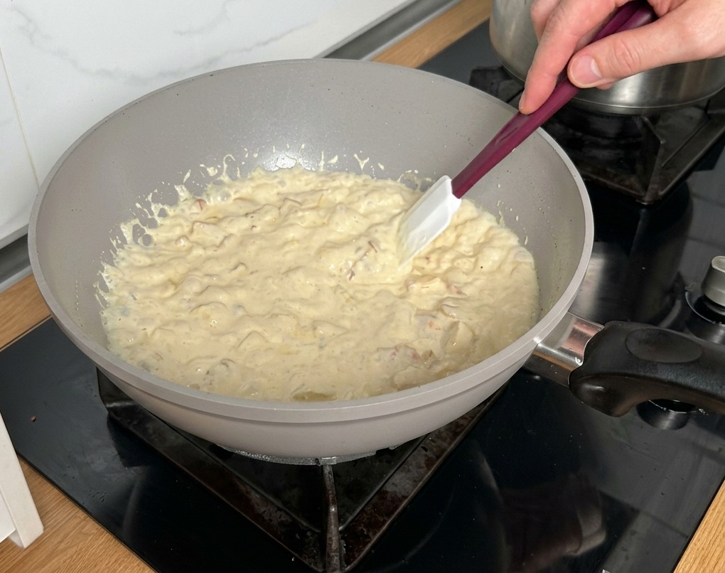A white spatula stirring a thick, bubbling cream sauce with bacon and mushrooms in a light-colored pan.