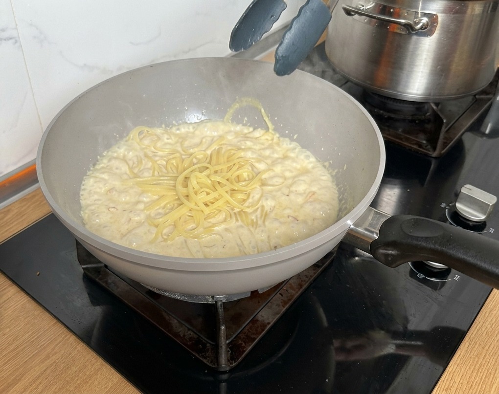 Metal tongs lifting cooked pasta into a pan filled with creamy bacon and mushroom sauce.