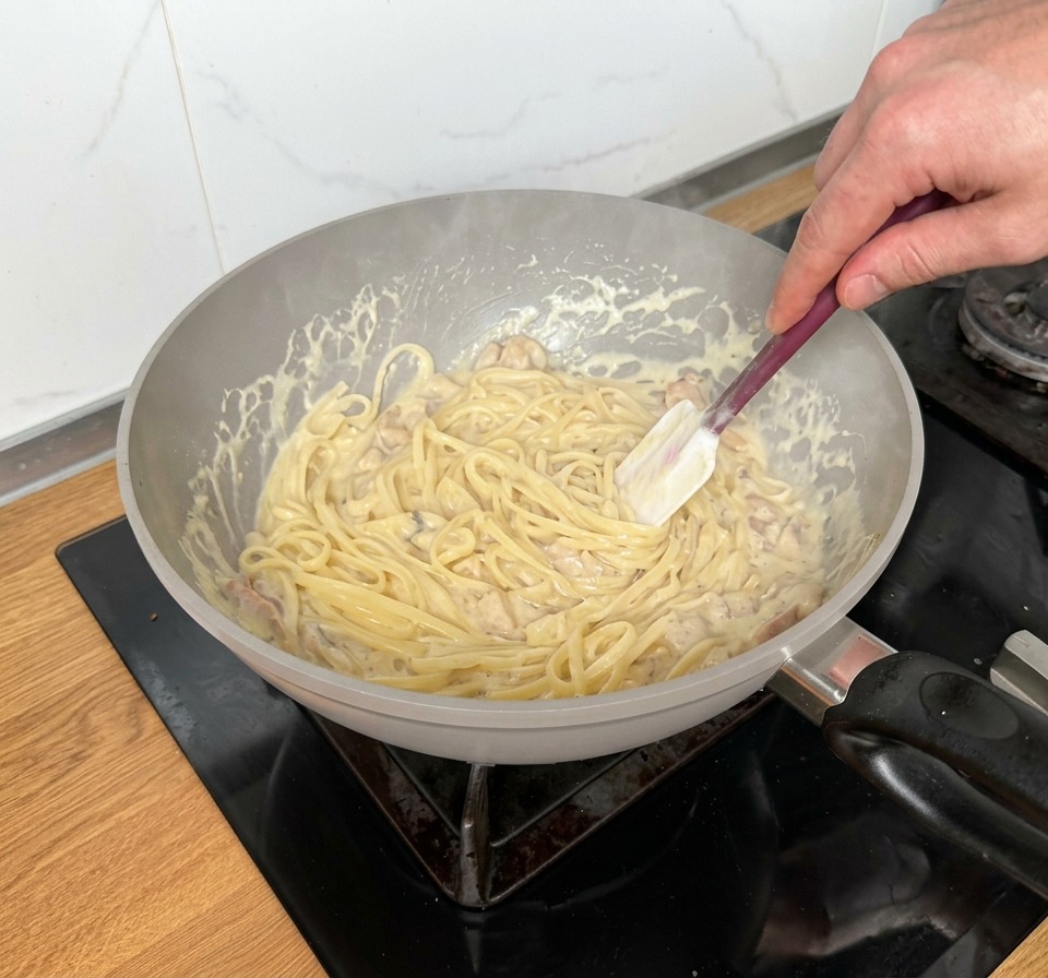 Tossing linguine pasta in a pan filled with a creamy, rich sauce, bacon strips, and quartered mushrooms.