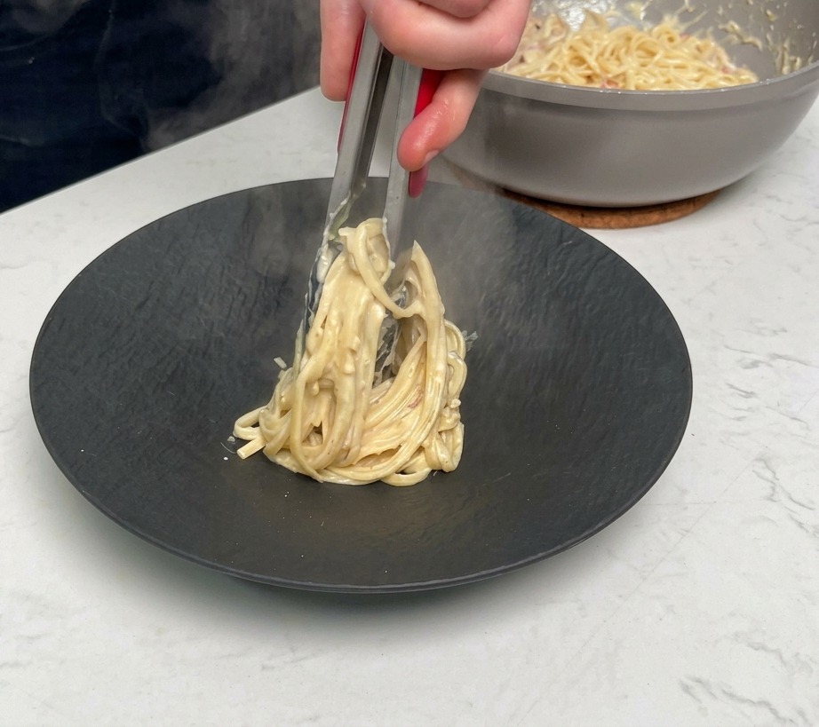 Using metal tongs to twirl and plate a serving of creamy bacon pasta onto a matte black dish.