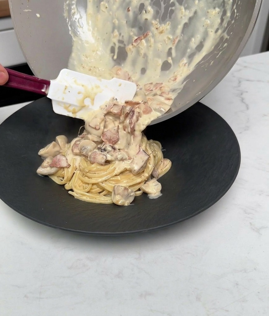 Pouring leftover creamy sauce, cooked mushrooms, and bacon from a pan over the plated pasta.
