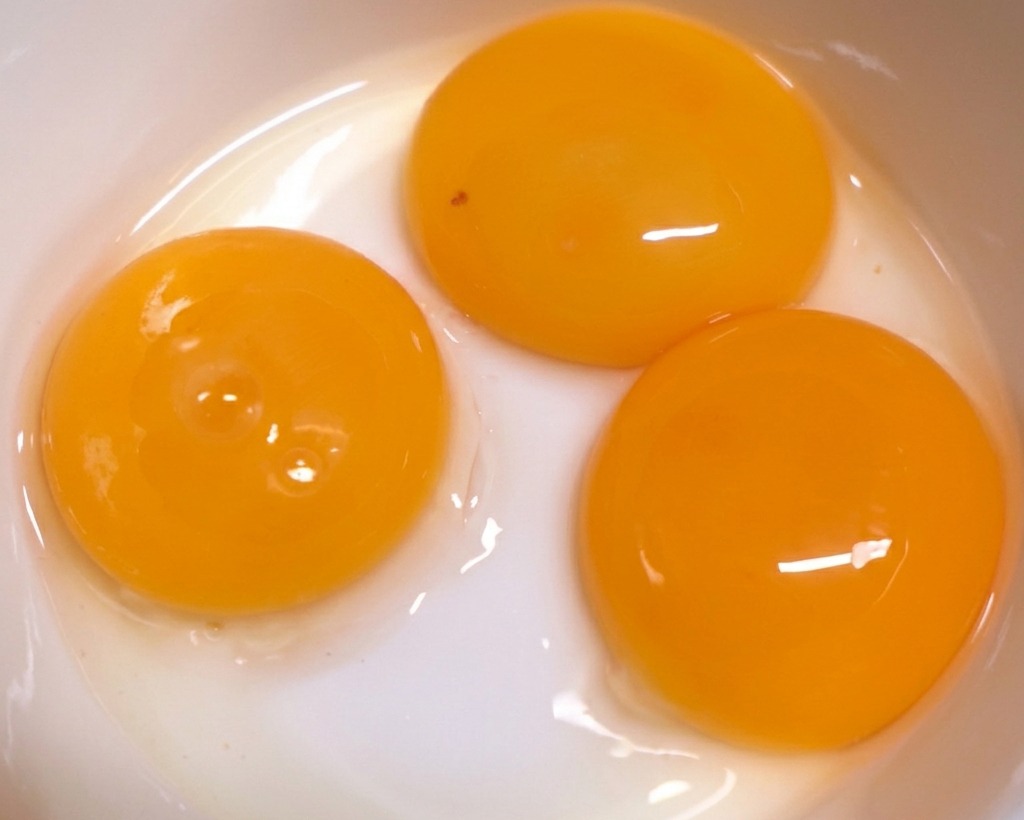 Three vibrant orange egg yolks sitting in a clean white ceramic bowl, ready for preparation.
