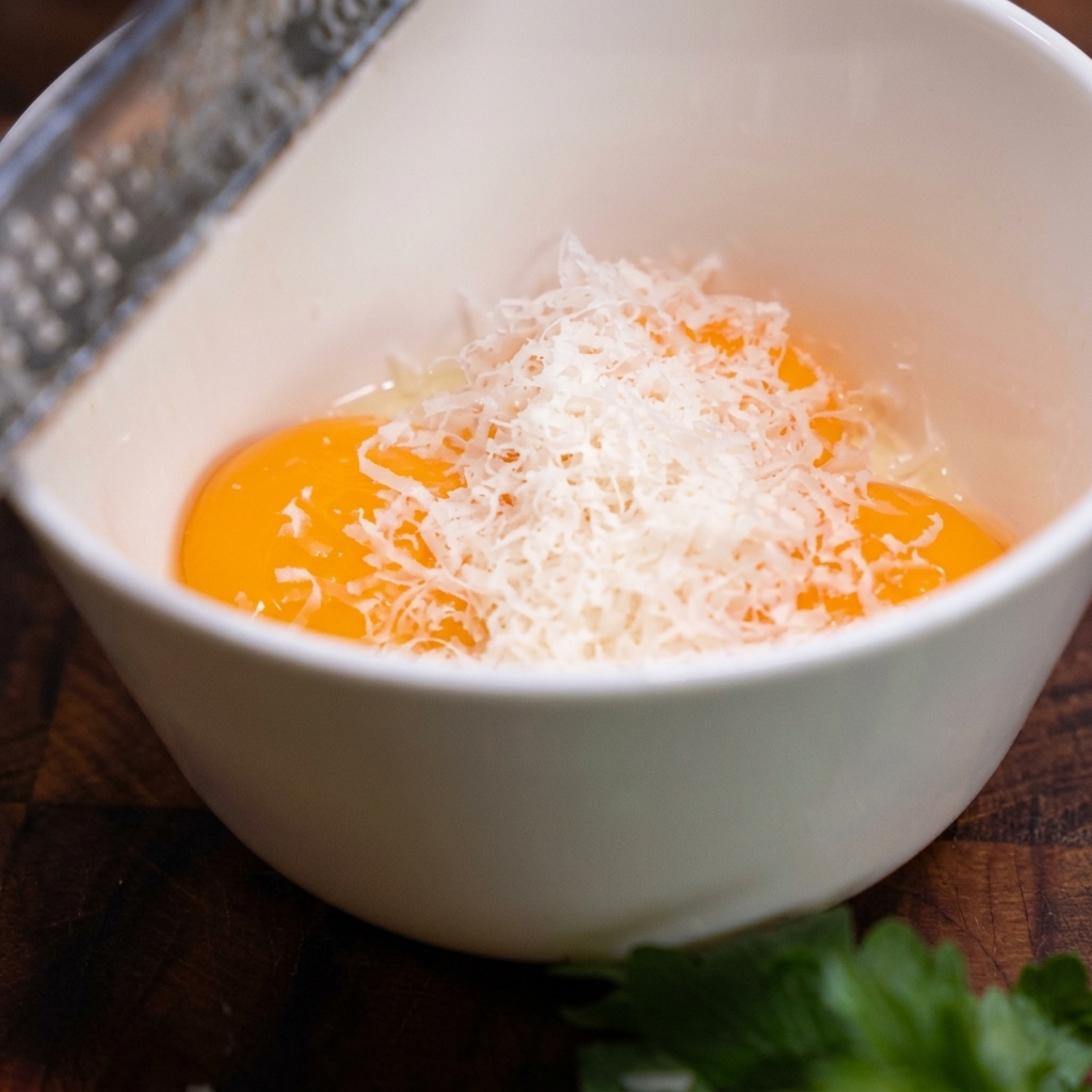 A metal grater shedding fine white curls of Parmesan cheese into a bowl containing three raw egg yolks.