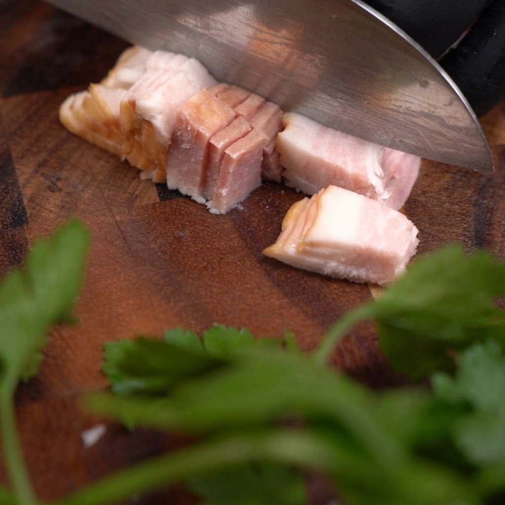A sharp knife cutting through a slab of raw bacon to create small, uniform cubes on a dark wooden surface.