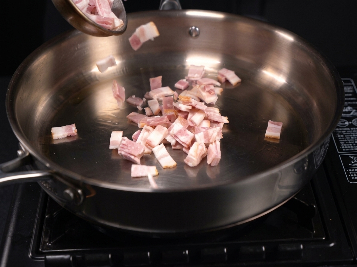 Small, chopped pieces of bacon sizzling and releasing fat in a stainless steel frying pan.