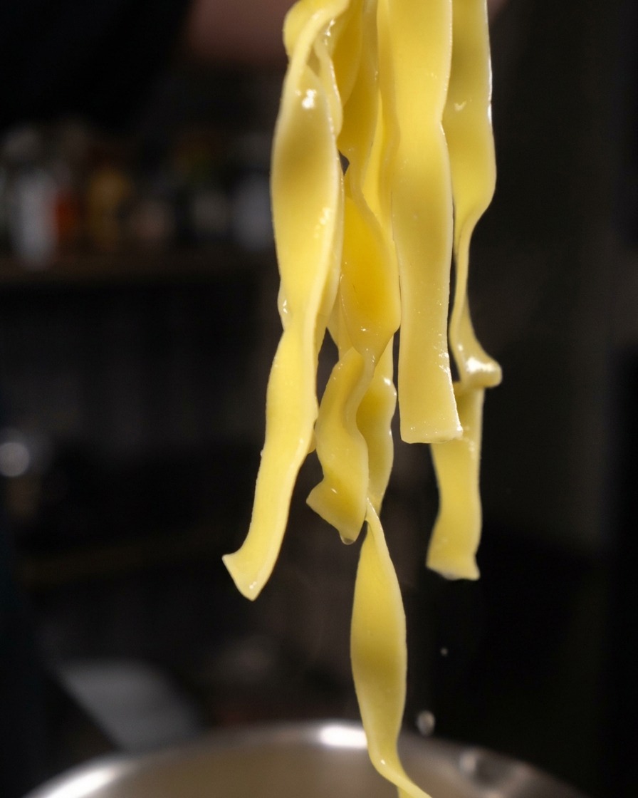 Long, thick strands of cooked yellow tagliatelle pasta hanging from tongs, dripping water.