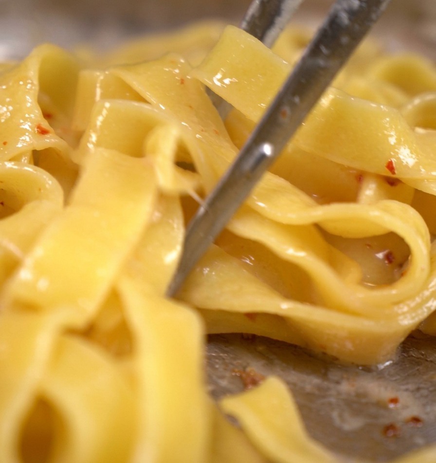 Tagliatelle pasta being tossed with metal tongs in a pan with bacon bits and simmering pasta water.
