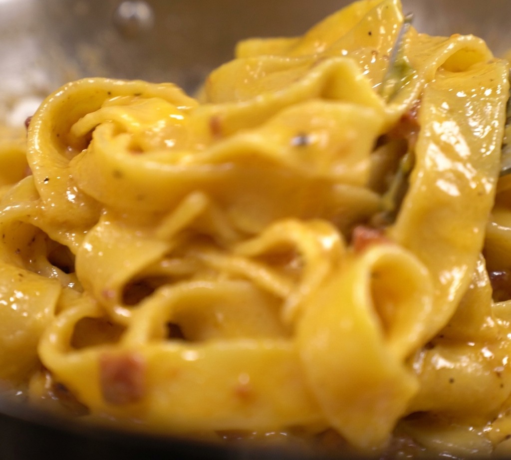 Tagliatelle pasta coated in a thick, vibrant yellow creamy egg and cheese sauce with visible bacon bits in a pan.