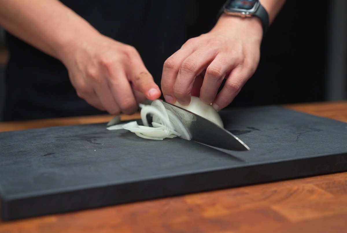 Hands holding a chefs knife, slicing a white onion into thin strips on a dark cutting board.