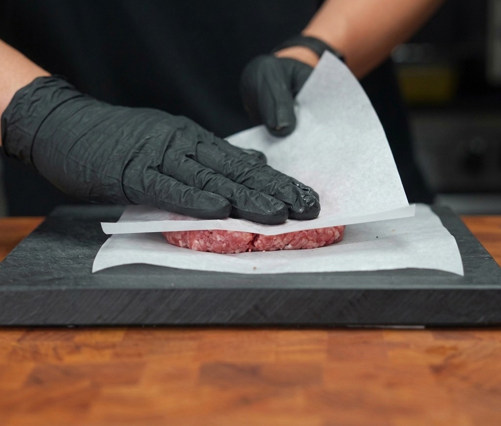 Gloved hands firmly pressing a raw beef patty flat between two sheets of white parchment paper on a cutting board.