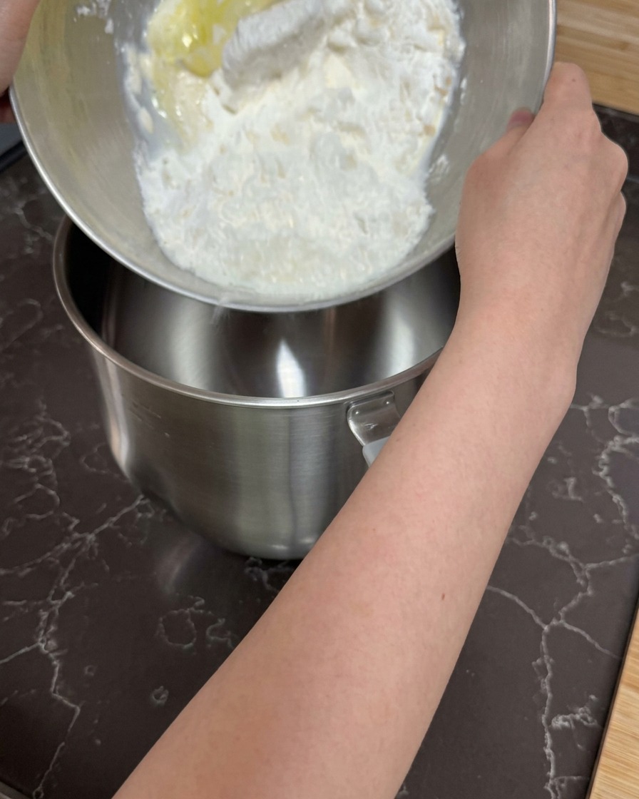 Hands pouring a shaggy mixture of flour and wet ingredients into the empty bowl of a stand mixer.
