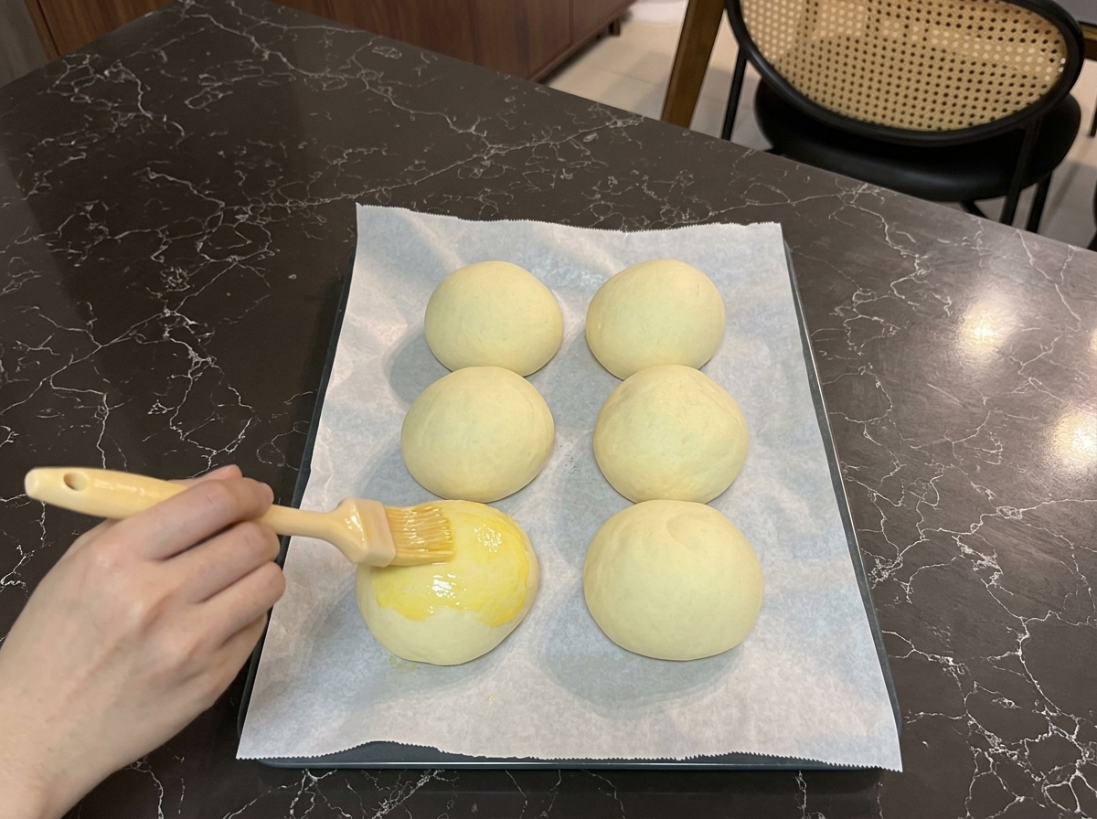 A hand using a pastry brush to coat the top of a proofed burger bun with a glossy yellow egg wash.