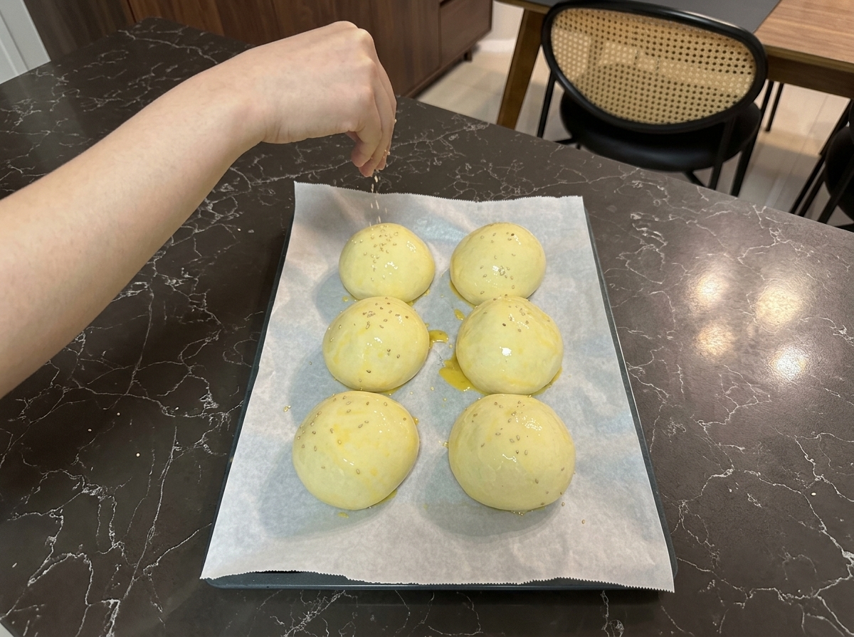 A hand sprinkling tiny white sesame seeds over egg-washed dough balls arranged on a baking sheet.