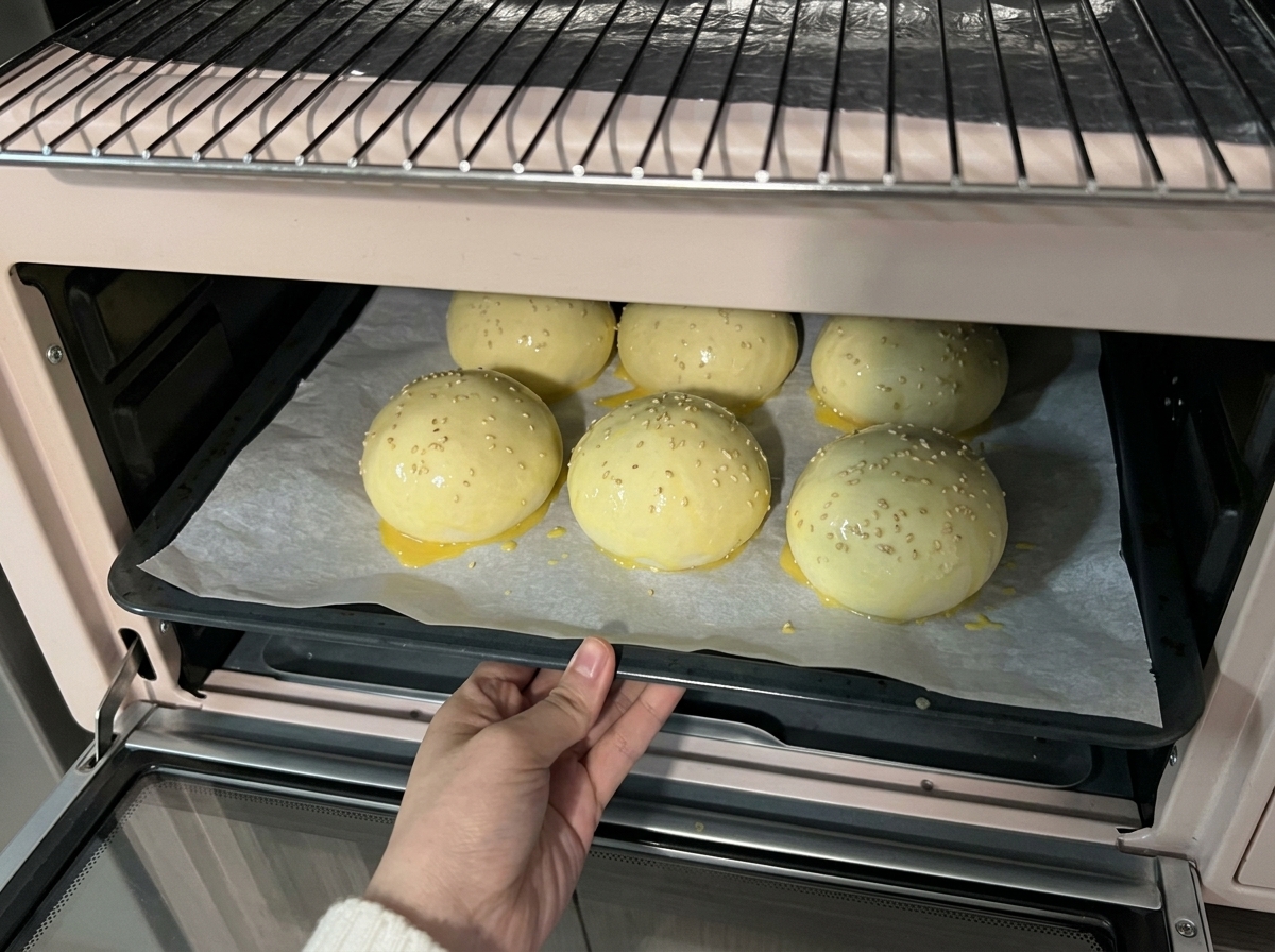 A tray of sesame-topped dough balls being slid into a light pink counter-top oven for baking.