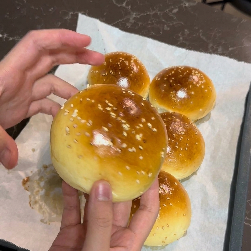 A persons hands gently holding a freshly baked, golden-brown burger bun sprinkled with white sesame seeds.