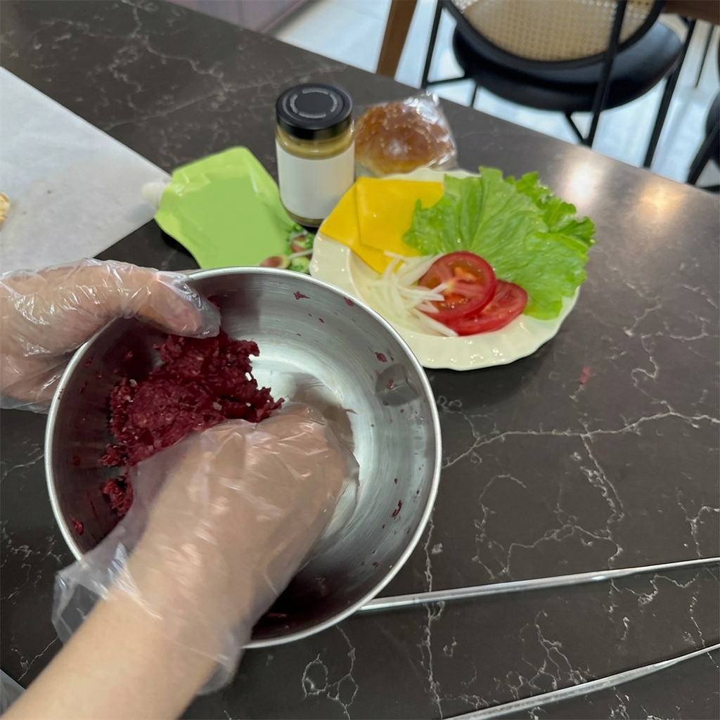 Hands wearing clear plastic gloves mixing raw, bright red ground beef inside a stainless steel mixing bowl.